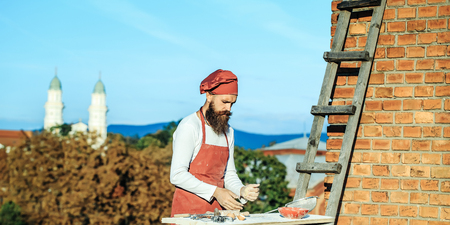 Man cook chef hipster with beard on handsome face in red hat and white uniform rolling dough with pin near ladder brick wall blue sky and green trees background outdoorの写真素材
