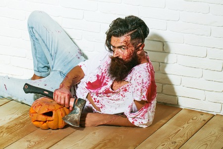 Bearded zombie man with beard vampire or bloody war soldier with wounds and red blood with halloween pumpkin and axe lies on wooden floorの写真素材