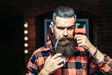 Handsome bearded man hipster with stylish haircut and beard holding scissors in red checkered shirt near mirror with serious face in beauty salon or barbershop as barberの写真素材