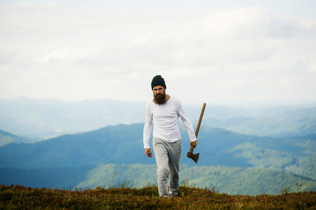 Handsome man bearded hipster with beard and moustache in hat walks with axe on mountain top on cloudy skyの写真素材