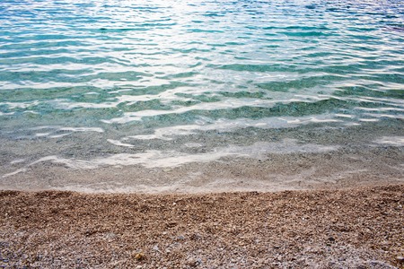 Blue sea or ocean clear shiny shallow water washes pebble stony shore beach on sunny summer dayの写真素材