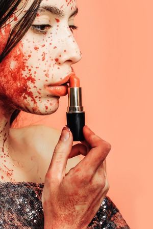 Halloween zombie girl or pretty young woman with brunette hair with wounds and blood puts red lipstick on lips on pink backgroundの写真素材