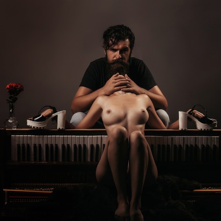 handsome bearded man with long beard on serious face near young woman or girl topless with sexy beautiful bare chest or breast on naked body and belly in panties near old wooden piano keyboardの写真素材