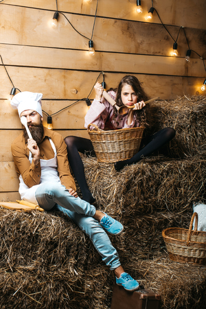 Funny man chef cook or baker with beard and moustache in hat toque and cute girl cookee teenager in apron sit on straw bales on rustic backgroundの写真素材