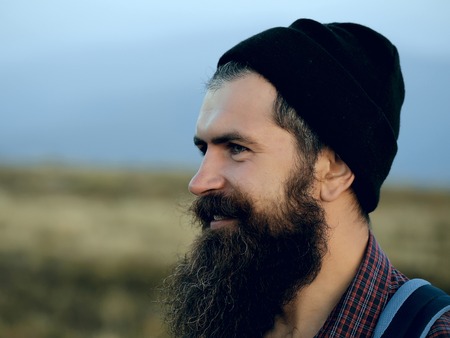 handsome bearded man with mustache and long lush beard on happy face in black hat with backpack on blurred background, close upの写真素材