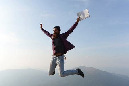 handsome bearded man with mustache and long lush beard on serious face with backpack and map jumping on blue sky backgroundの写真素材