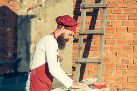 Chef man hipster with long beard on handsome face in red hat and apron white uniform rolling dough...