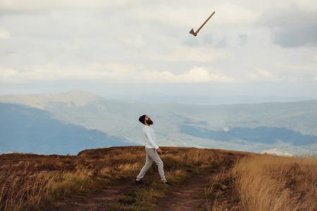 Handsome man bearded hipster with beard and moustache in hat throws axe on mountain top on cloudy skyの写真素材
