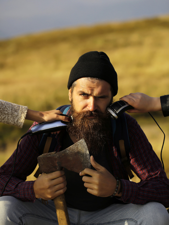 handsome bearded man with mustache and long lush beard on face sit with axe and two female hands shave him on mountain top on blurred backgroundの写真素材