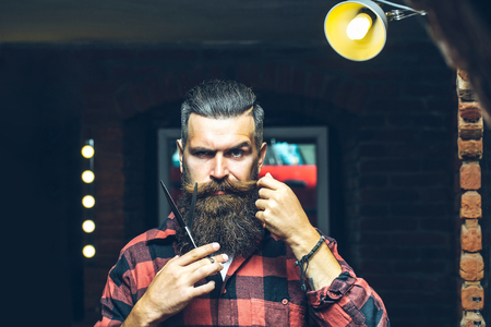 Handsome bearded man hipster with stylish haircut and beard holding scissors in red checkered shirt near mirror with serious face in beauty salon or barbershop as barber near brick wallの写真素材
