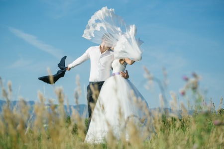 Handsome groom man shirt sleeve holds beautiful bride woman in long white wedding dress with flying veil in summer field on blue sky backgroundの写真素材