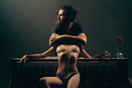 handsome bearded man with long beard on serious face near young woman or girl topless with sexy beautiful bare chest or breast on naked body and belly in panties near old wooden piano keyboardの写真素材