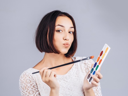 pretty sexy cute woman or girl with short brunette hair in white tracery blouse with colorful paint and long paintbrush on gray studio backgroundの写真素材