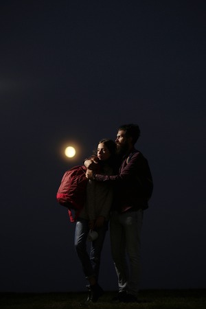 couple of strong man hipster with long beard and moustache in checkered shirt and sexy woman or cute girl with backpack on twilight embracing on darknessの写真素材