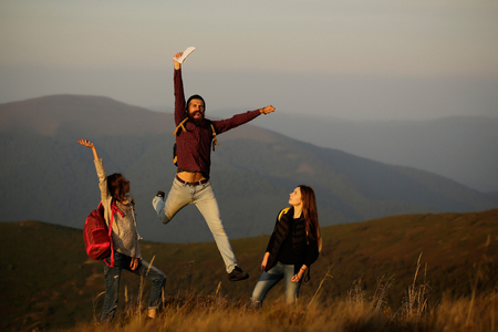 handsome bearded man in checkered shirt with pretty young cute girls or women walking shouting and jumping with map on mountain top on sunny dayの写真素材
