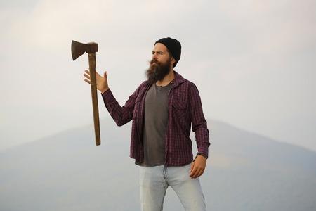 young handsome bearded hipster man in checkered shirt with long beard holding axe or ax on mountain top on natural background with skyの写真素材