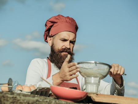 Man chef cook hipster with handsome bearded face in hat and white and red uniform bolting flour with...