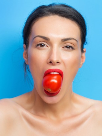 young cute woman or girl portrait with red tomato in sexy lips on pretty face and brunette hair has fashionable makeup on blue backgroundの写真素材