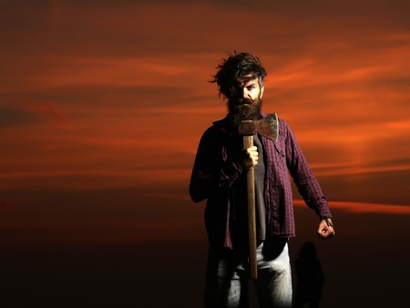 Strong man lumberjack hipster with long beard and moustache in checkered shirt with axe in hand on twilight on darkness backgroundの写真素材