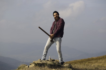 bearded man hipster with long beard and moustache in checkered shirt with axe in hand standing and smiling on stone on mountain top on sunny dayの写真素材