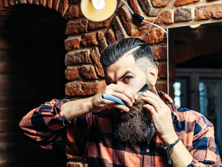 Handsome bearded man hipster with stylish haircut and beard holding old fashion razor in red checkered shirt with serious face in beauty salon or barbershop as barber near brick wallの写真素材