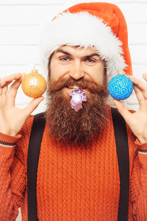 young handsome smiling bearded santa claus man with long beard in red sweater and new year hat holds decorative christmas or xmas balls on white brick wall backgroundの写真素材
