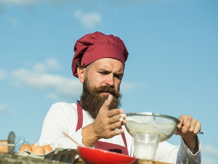 Man chef cook hipster with handsome bearded face in hat and white and red uniform bolting flour with sieve on blue sky background outdoorの写真素材