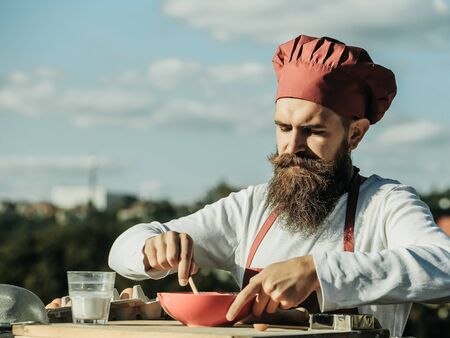 Man cook chef hipster with bearded handsome face in red uniform and hat mixing up ingredients with spoon in bowl on blue sky and roofs background outdoorの写真素材