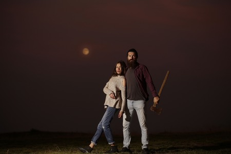 Young couple of pretty girl and bearded man hipster with beard and axe on mountain top at night on moon lightの写真素材