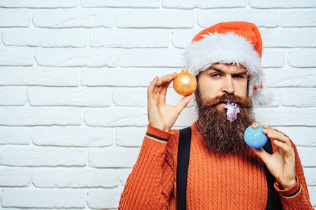 young handsome surprised bearded santa claus man with long beard in red sweater and new year hat holds decorative christmas or xmas balls on white brick wall background, copy spaceの写真素材