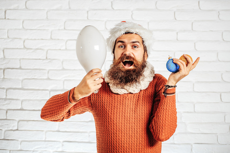 young handsome bearded santa claus man with long beard in red sweater and new year hat holds decorative christmas or xmas balls and big lamp on white brick wall backgroundの写真素材