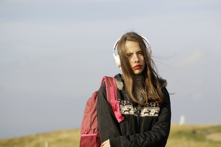Pretty girl young beautiful woman traveler with headphones and red backpack stands on mountain top...