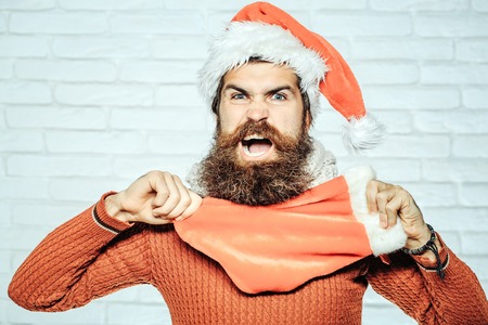 young handsome angry bearded santa claus man with long beard in red sweater and new year hat holds decorative christmas or xmas stocking or boot on white brick wall backgroundの写真素材