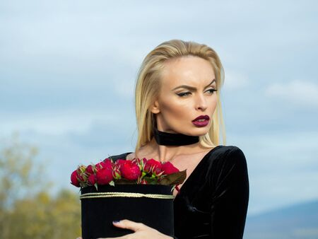 Pretty girl beautiful blond woman in sexy black dress with flowers in box outdoors on blue skyの写真素材