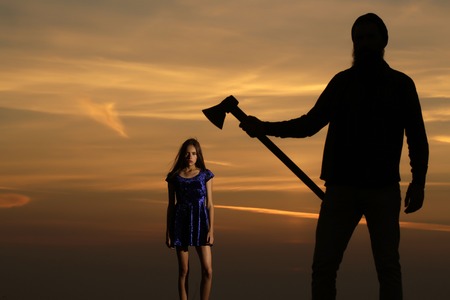 Dark silhouette of man with axe and pretty girl young beautiful woman stand on mountain top on sunset skyの写真素材