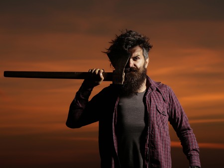 Brutal man bearded hipster with beard and moustache stands with axe on dark sunset skyの写真素材