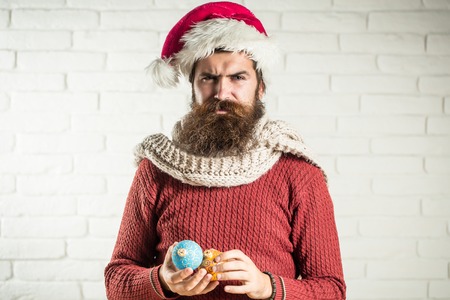 young handsome bearded santa claus man with long beard in red sweater and new year hat holds decorative christmas or xmas balls on white brick wall backgroundの写真素材
