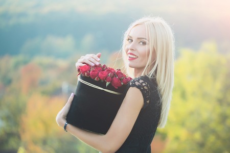 Pretty cute girl or beautiful blond woman in sexy black dress with red rose flowers in box outdoor on natural backgroundの写真素材