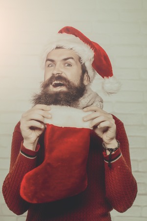 young handsome bearded santa claus man with long beard in red sweater and new year hat holds decorative christmas or xmas stocking or boot on white brick wall backgroundの写真素材