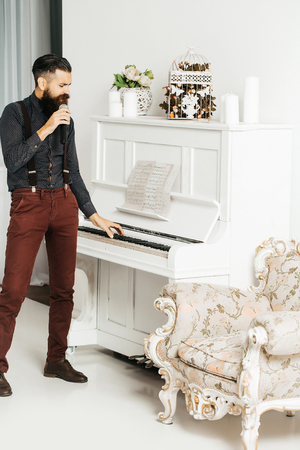 Handsome bearded young man in fashionable shirt and pants near luxurious baroque decorated with flowers armchair playing white piano and singing in mic or microphoneの写真素材