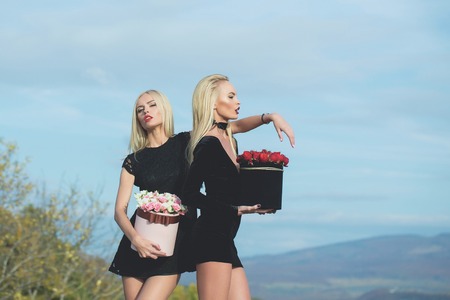 Pretty cute girls or beautiful blond women twins in sexy black dress with red and white rose flowers in box outdoor on natural background with blue cloudy skyの写真素材