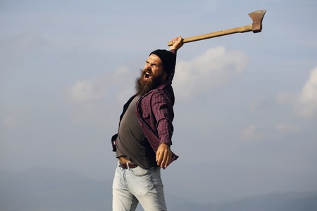 young handsome bearded hipster man in checkered shirt with long beard holding axe or ax on mountain...