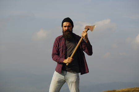 young handsome bearded hipster man in checkered shirt with long beard holding axe or ax on mountain top on natural background with skyの写真素材