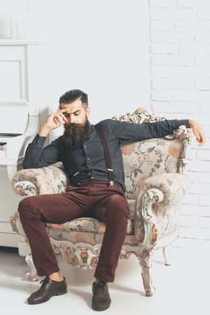Handsome bearded young man in fashionable shirt and pants sit on luxurious baroque decorated with flowers armchair near white pianoの写真素材