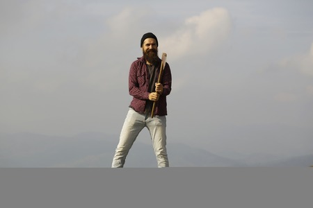 young handsome bearded hipster man in checkered shirt with long beard holding axe or ax on mountain...