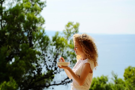 Pretty cute woman or sexy girl with curly blond hair stands with plate and spoon of morning breakfast eating food on balcony on summer day on natural backgroundの写真素材