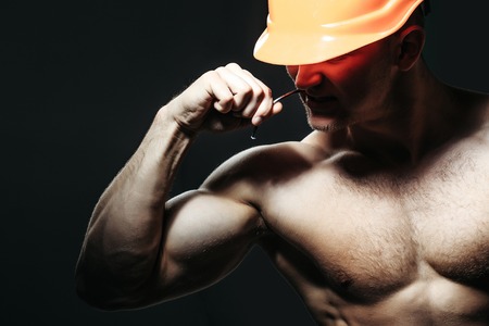 young handsome macho man builder with sexy muscular athletic strong body has bare chest and strong hands in orange hard hat or helmet holds spike or stud on black backgroundの写真素材