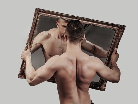 young handsome macho man with sexy muscular athletic body with bare torso and strong back holds big mirror on grey backgroundの写真素材