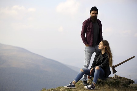 Couple of handsome bearded hipster in checkered shirt and pretty cute sexy young fashion girl or woman sitting on stone with ax on mountain top on natural backgroundの写真素材
