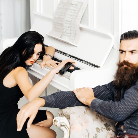 Pretty sexy young girl with long dark hair red lipstick on in black dress with microphone in hand sits at white piano and handsome bearded man in grey dotted shirt sits near in luxurious armchairの写真素材